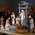 A carved nativity scene set out on a wood table laid with a linen cloth and lit candles in the background.