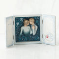 Open light wooden book with three parts - center is blue sky with two carved in faceless boy and girls hugging one another