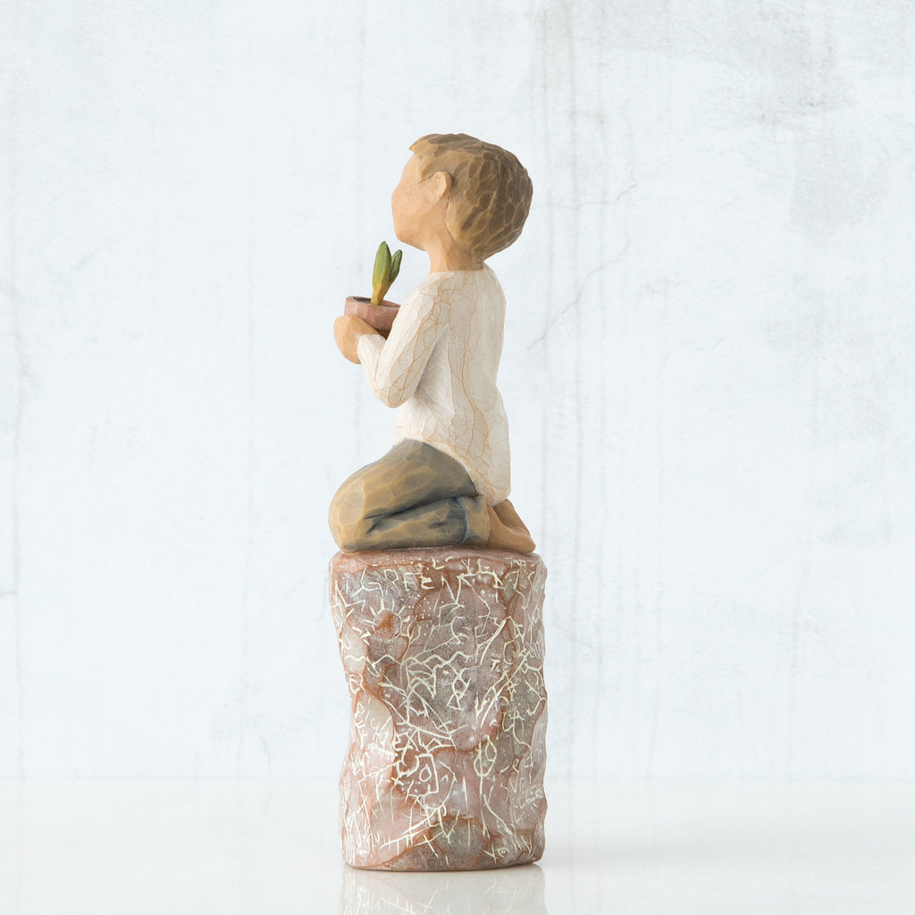 Right side view of boy with brown hair sitting on a stump while holding a small potted plant to his chest