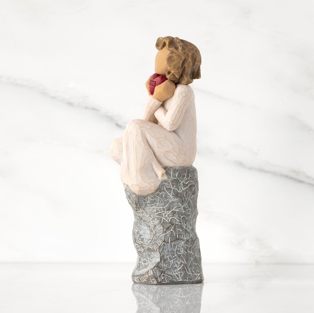 Facing left: figure in cream dress, seated on gray rock, knees drawn up to chest, holding red heart engraved with year 2023