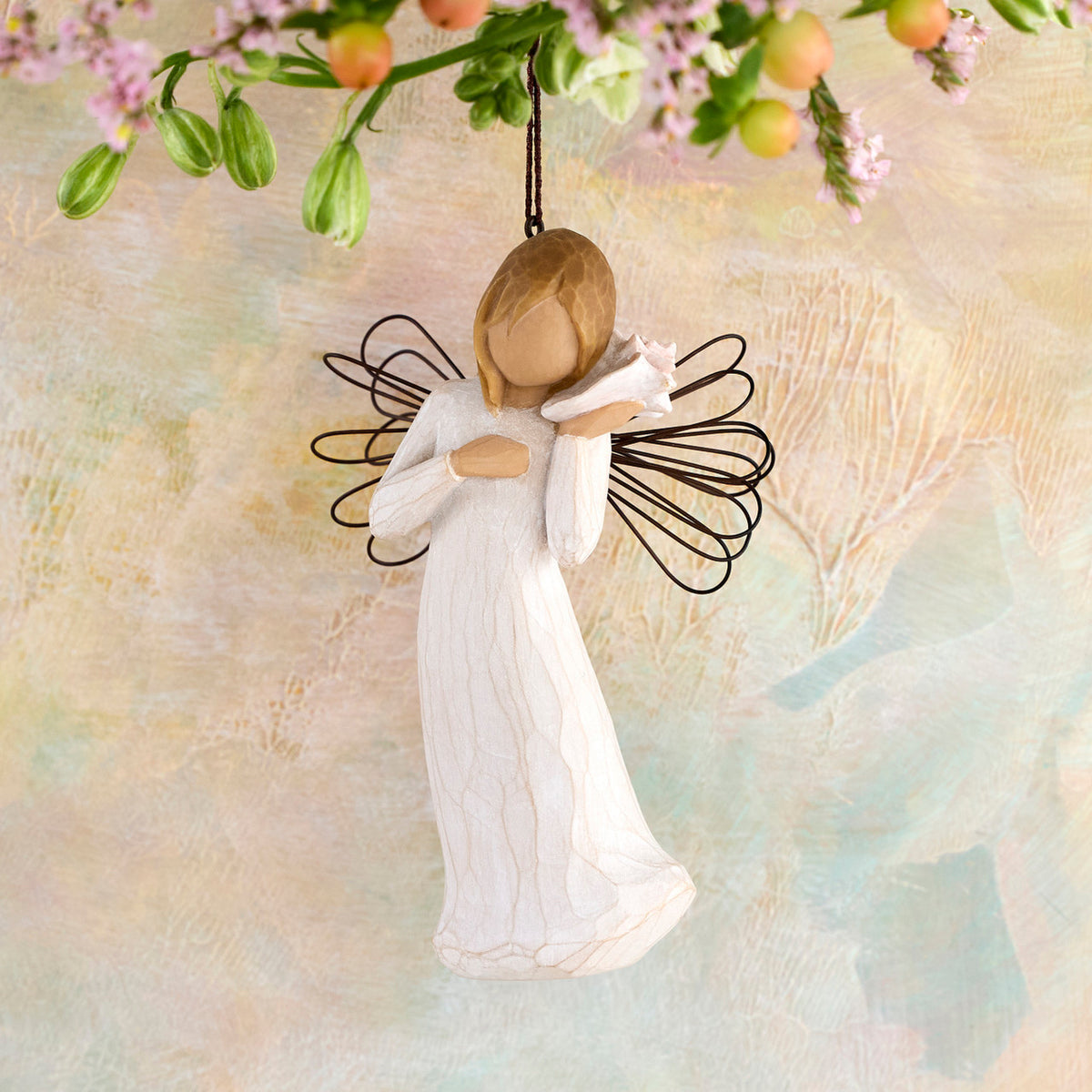 Willow Tree Thinking of You Ornament, 4.5”h hand-painted resin angel figure in cream dress with wire wings, holding pink conch shell to ear, brown elastic cord, hanging in front of peach painting