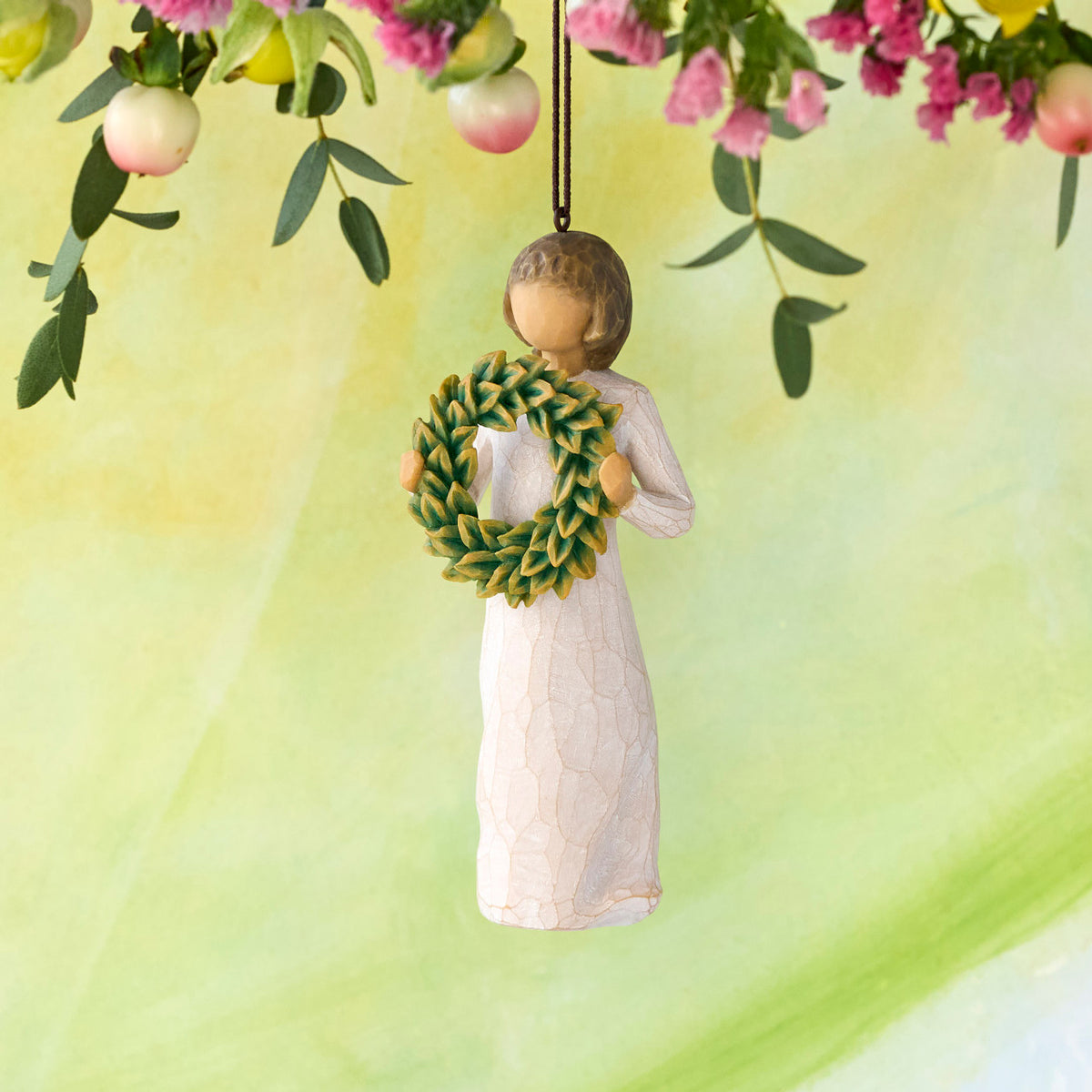 Willow Tree Magnolia Ornament, 4.25”h hand-painted resin figure in cream dress, holding green magnolia leaf wreath, brown elastic cord, hanging in front of pale green painting