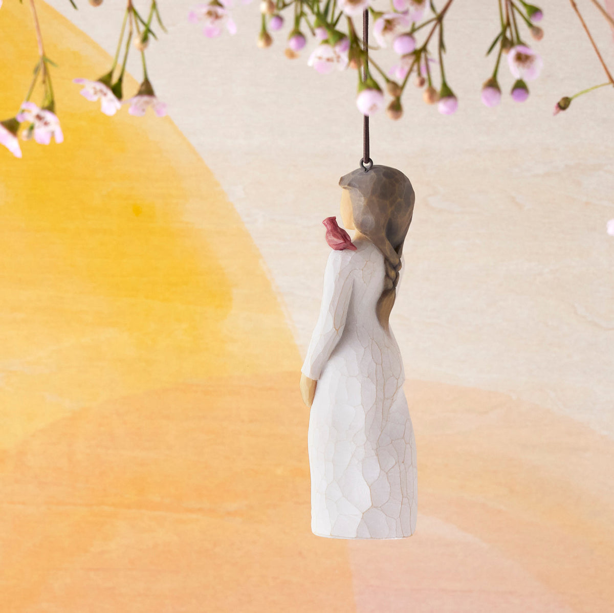 Willow Tree Messenger Ornament, side view of 4”h hand-painted resin figure in cream dress with red cardinal on shoulder, brown elastic cord, hanging in front of orange painting